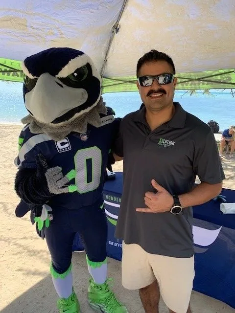 Blitz, the Seattle Seahawks mascot, posing with a man in an EVlution Energy polo shirt under a canopy at a beach cleanup event
